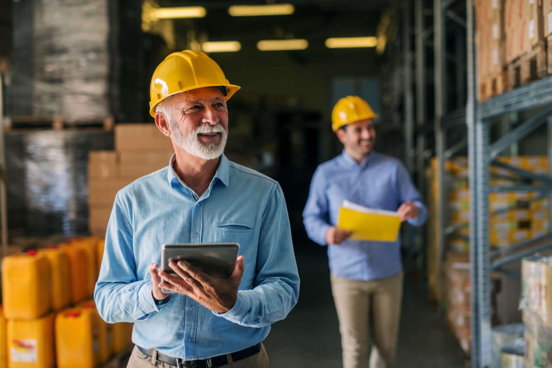 two professional construction leaders, one older with a tablet in the focused foreground and one younger in the blurred background holding a folder