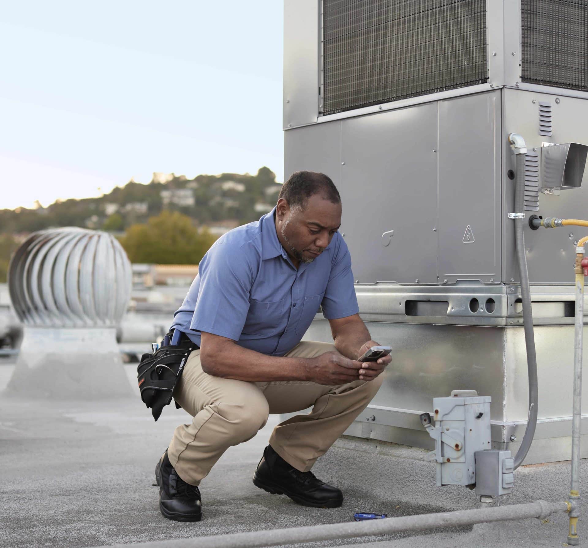 service provider crouching near industrial rooftop unit reviewing information on their smartphone