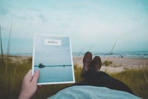 person pictured laying down with feet in front, looking out over a beach with magazine in hand