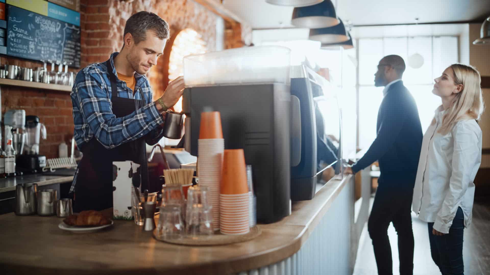 Male Barista in Checkered Shirt is Making a Latte for a Customer in a Coffee Shop