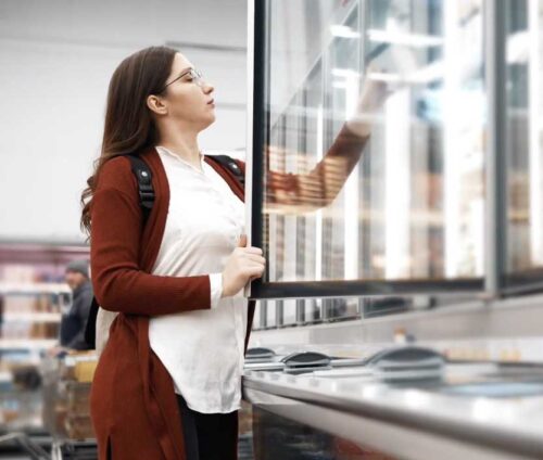 middle aged woman in maroon cardigan picking a refrigerated product from the cooler in a retail store