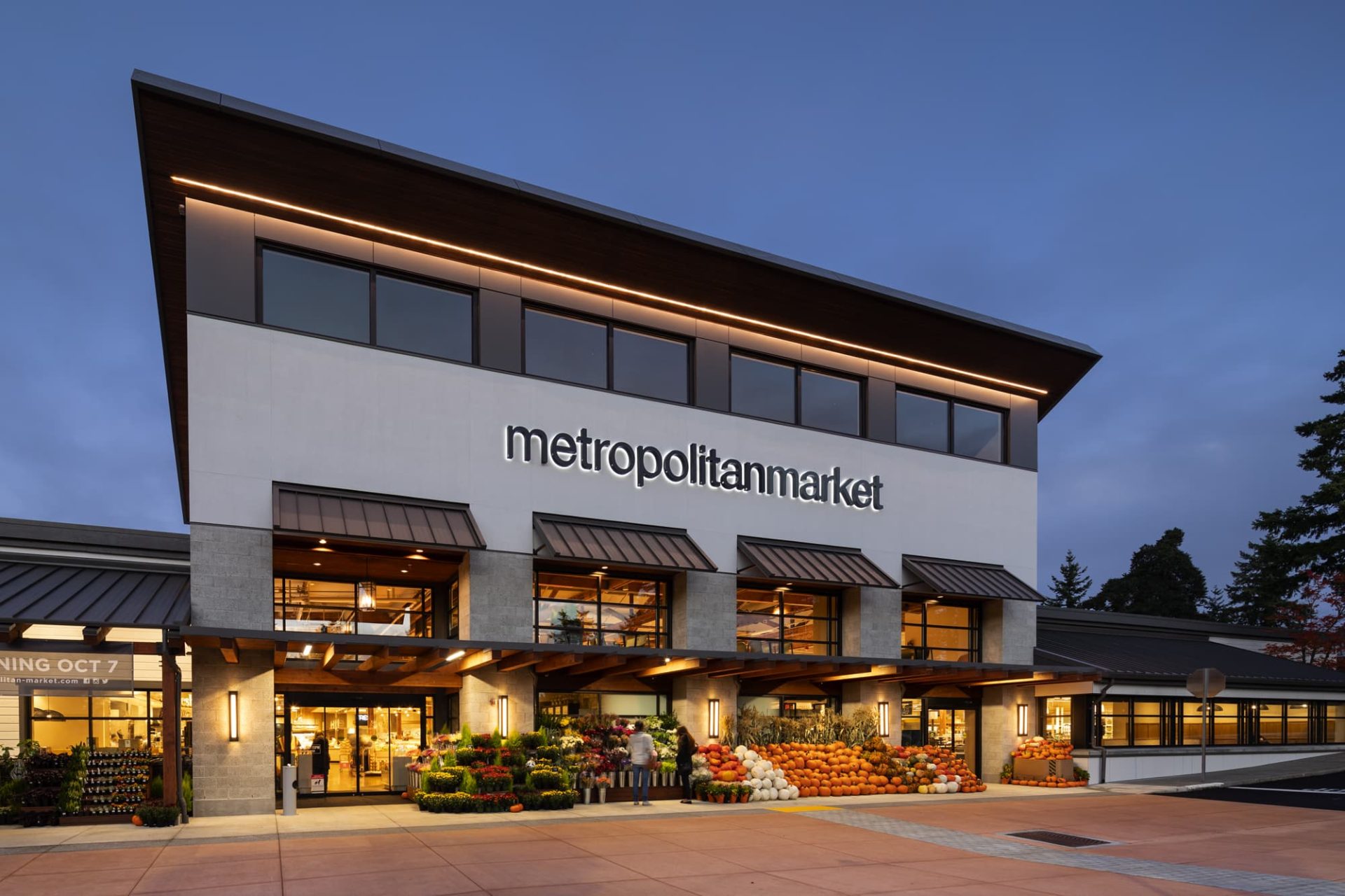 metropolitan market storefront with plants, pumpkins, and passerby in an evening setting